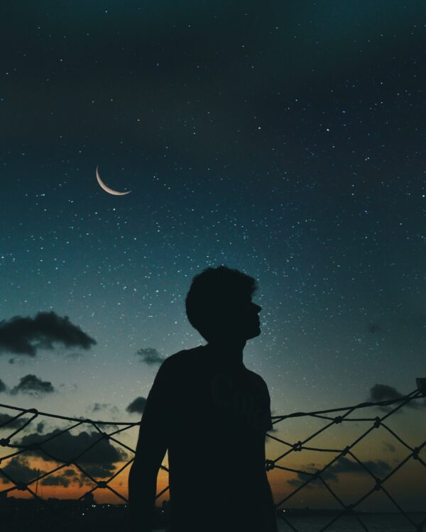 A lone silhouette stands beneath a crescent moon and starry sky, evoking a sense of solitude and wonder.