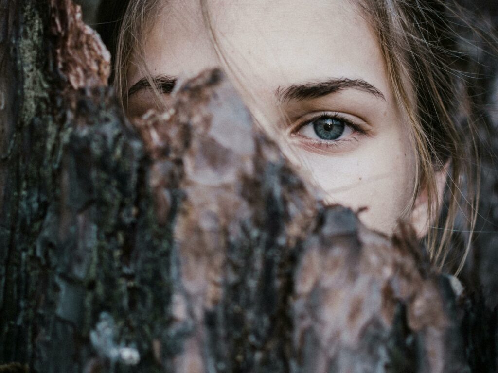 A mysterious close-up of a woman's blue eye peeking from behind tree bark in an outdoor natural setting.