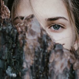 A mysterious close-up of a woman's blue eye peeking from behind tree bark in an outdoor natural setting.