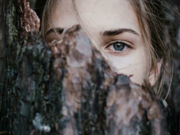 A mysterious close-up of a woman's blue eye peeking from behind tree bark in an outdoor natural setting.