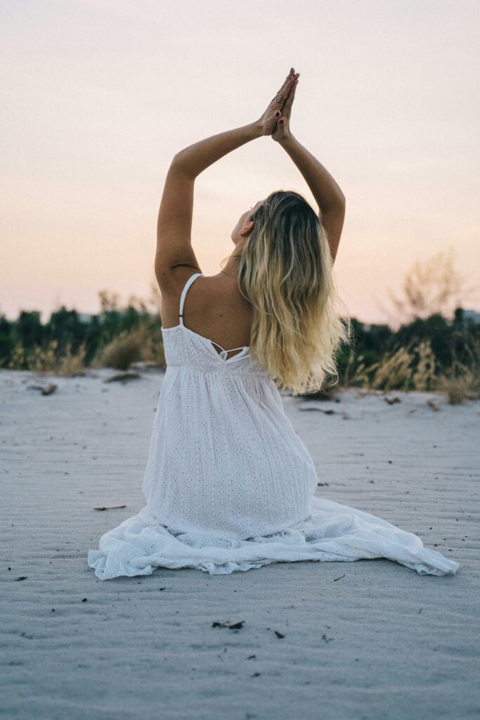 Woman in a white dress meditating on a serene beach during sunset, enhancing calmness and mindfulness.