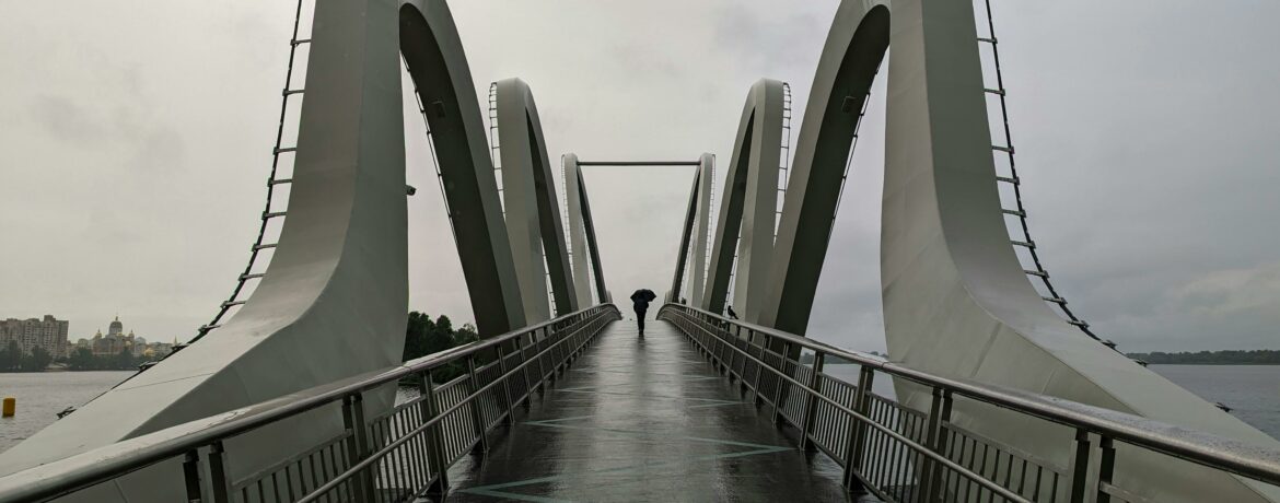 A solitary figure walks across an iconic modern bridge in Kyiv, Ukraine, against a moody sky.
