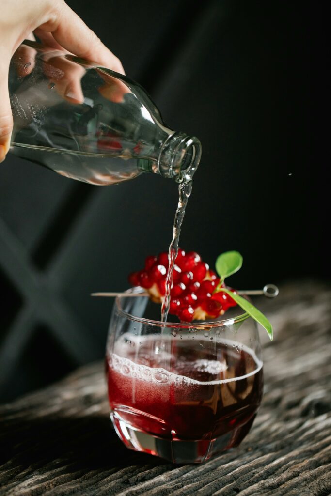 An elegant pour of sparkling water into a pomegranate garnished glass on a dark wooden surface.