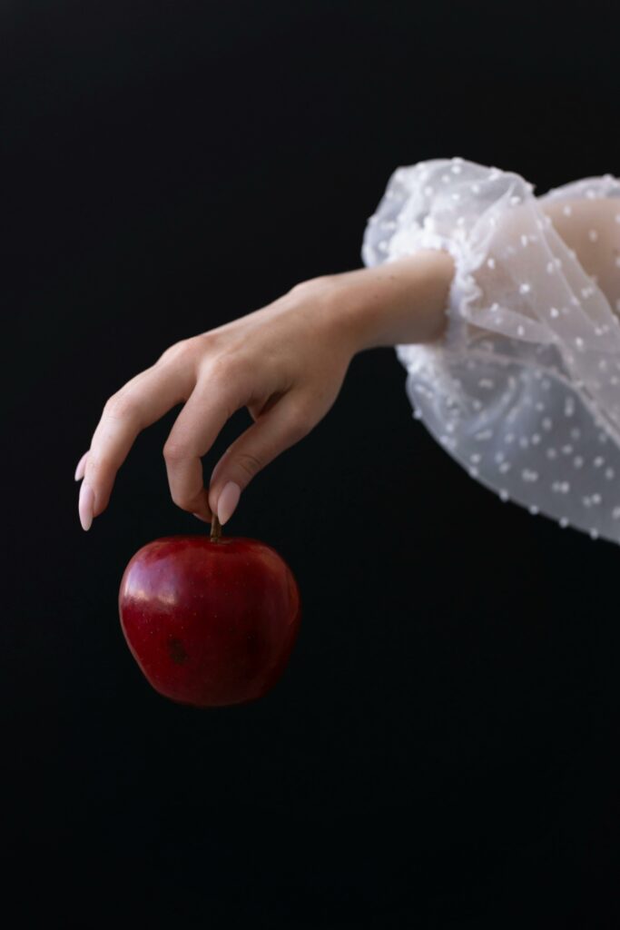 Delicate hand in white sleeve holding a red apple against a dark background, symbolizing temptation and elegance.
